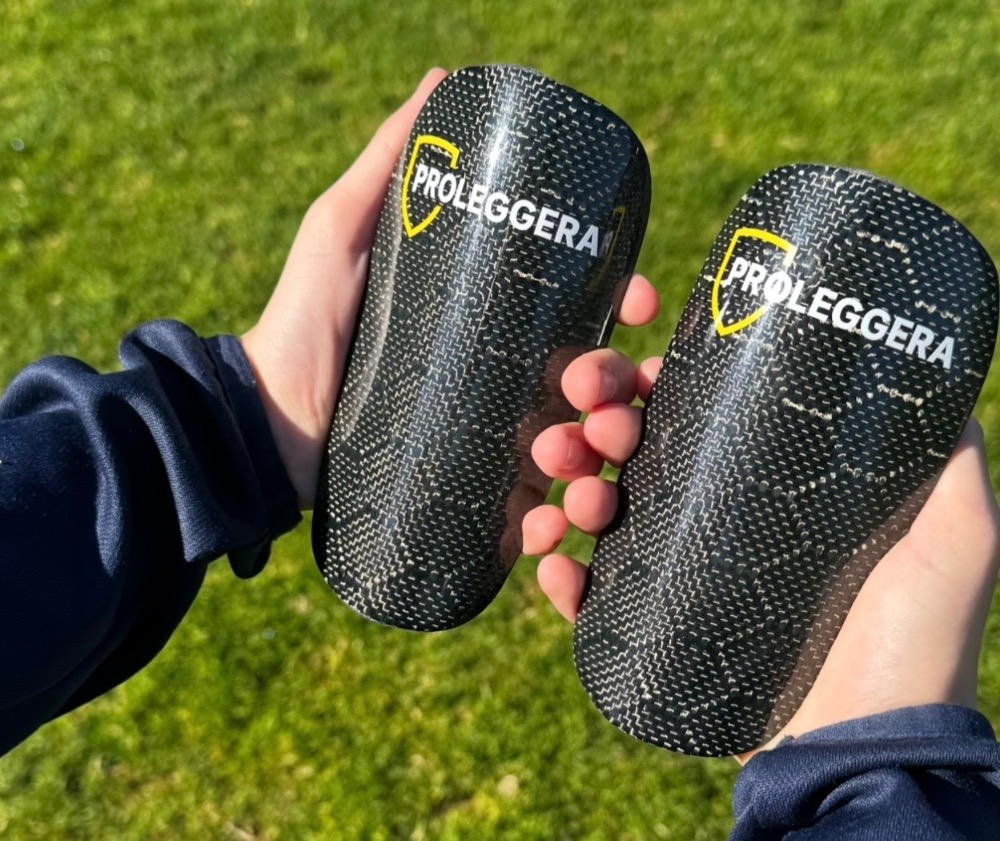 Fotbalové chrániče s logem PROLEGGERA vyrobené z karbonu, ve dlaních mladého fotbalisty nad zelenou trávou fotbalového hřiště.
