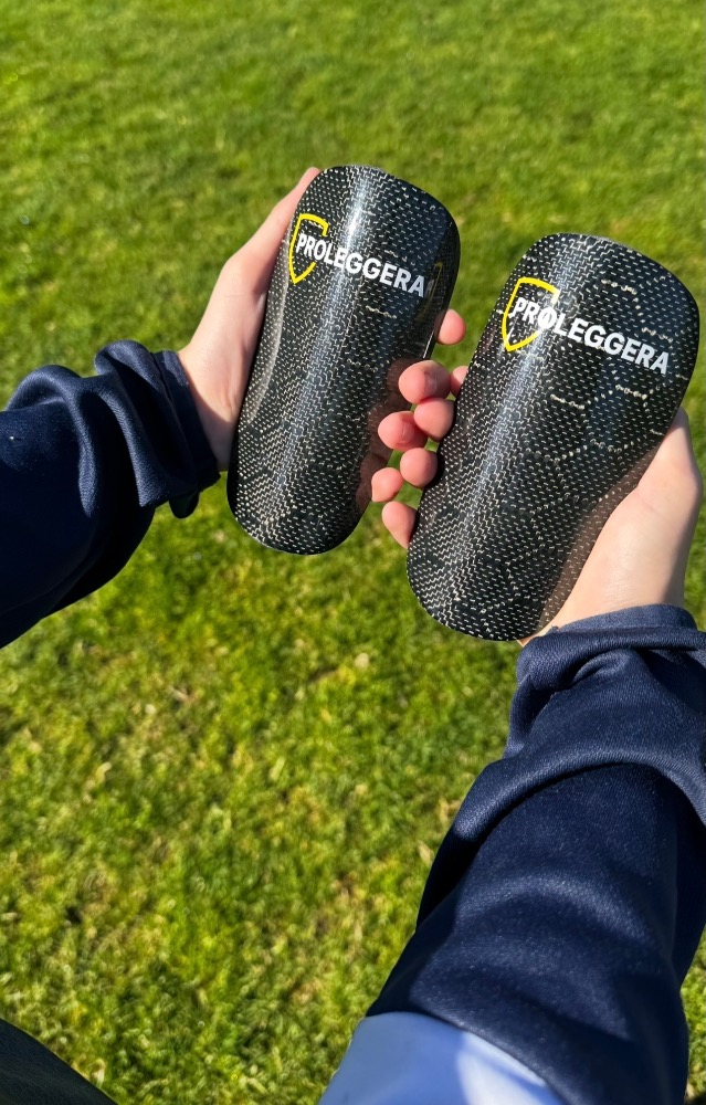 Due mani tengono parastinchi da calcio in carbonio con logo PROLEGGERA sopra l’erba verde di un campo da calcio.